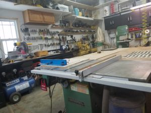 Picture of the workshop in a one car garage.  The table saw and two workbenches are in the center with more counters and tools setting along the outside walls.